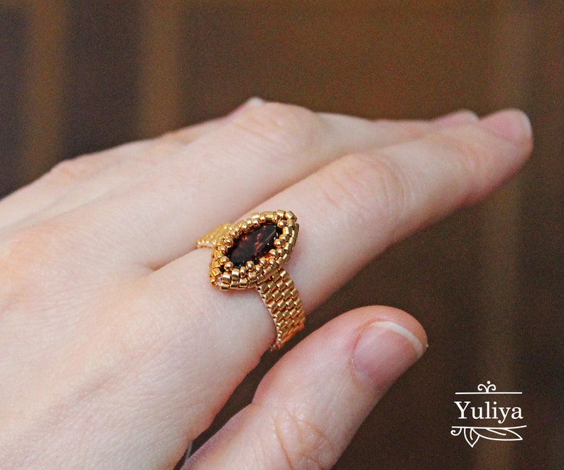 K&ouml;nnte beinhalten: Ein goldener Ring mit einem braunen, markisenf&ouml;rmigen Edelstein, der in einem Perlmuster gefasst ist.