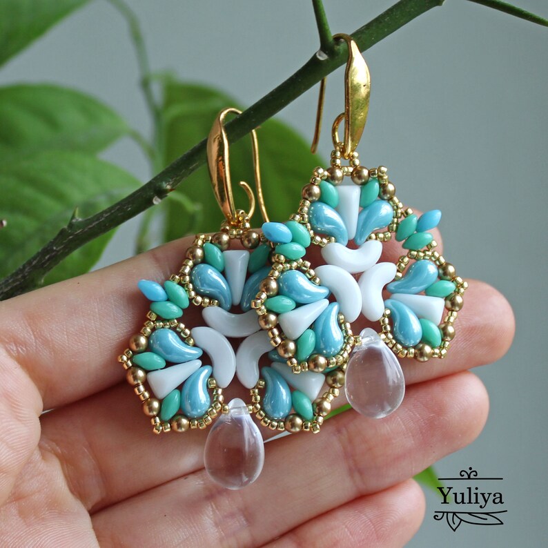 May include: Gold dangle earrings with a geometric design featuring white, turquoise, and light blue beads. The earrings have clear crystal drops.