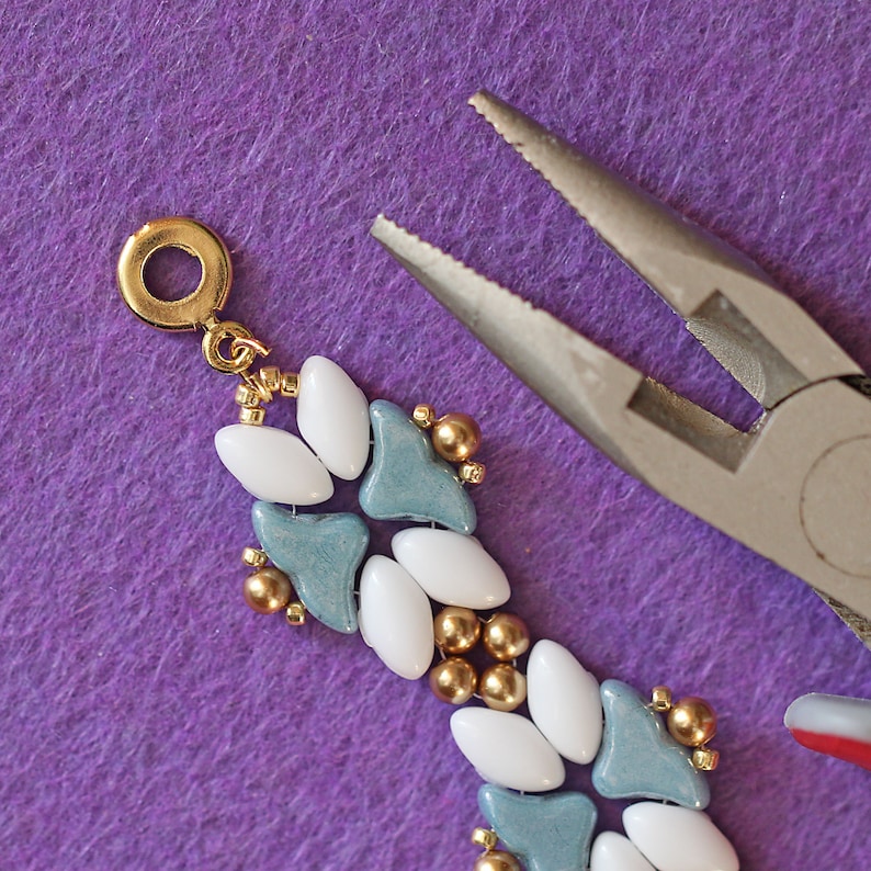 May include: Close-up of a beaded bracelet with white and blue beads and gold accents. The bracelet is being held by a pair of pliers.