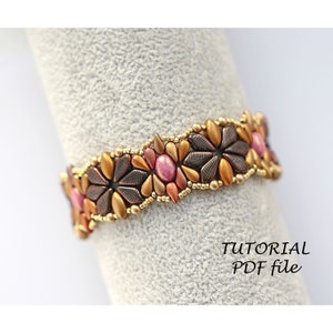 Pode incluir: Um bracelete de contas com um design floral em tons de ouro, cobre e rosa. O bracelete é feito com contas de sementes e apresenta um padrão repetitivo de flores. O bracelete é exibido em uma superfície branca. TUTORIAL PDF file.