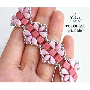 Könnte beinhalten: Ein rosa und silbernes Perlenarmband mit geometrischem Muster. Das Armband ist aus Rocailles gefertigt und weist ein sich wiederholendes Muster aus Quadraten und Tropfenformen auf. Das Armband wird in einer Hand gehalten, und der Text "Yuliya TUTORIAL PDF file" ist in der oberen rechten Ecke des Bildes sichtbar.