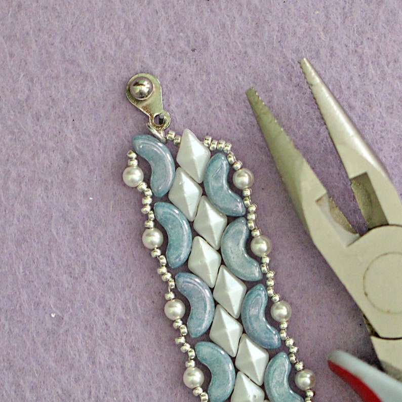 May include: Close-up of a silver clasp attached to a beaded bracelet with blue and white beads. A pair of pliers is in the background.