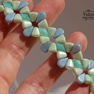 Peut inclure: Un bracelet de perles avec un motif r&eacute;p&eacute;titif de perles en forme de goutte d'eau dans des tons de bleu et de vert. Les perles sont dispos&eacute;es en zigzag, cr&eacute;ant un design unique. Le bracelet est tenu dans une main.