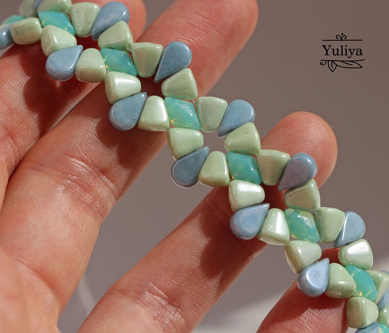 Peut inclure: Un bracelet de perles avec un motif r&eacute;p&eacute;titif de perles en forme de goutte d'eau dans des tons de bleu et de vert. Les perles sont dispos&eacute;es en zigzag, cr&eacute;ant un design unique. Le bracelet est tenu dans une main.