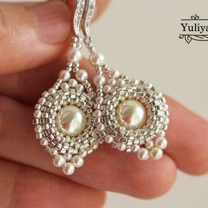 Może przedstawiać: Srebrne kolczyki z białymi perłami w okrągłym, koralikowym wzorze. Kolczyki mają zwisającą perłową kroplę.