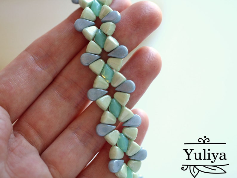 Peut inclure: Gros plan sur un bracelet de perles avec un motif r&eacute;p&eacute;titif de perles en forme de goutte d'eau, de couleur bleu clair, vert menthe et blanc cass&eacute;. Le bracelet est tenu dans une main, mettant en valeur le design et la palette de couleurs. C'est un bijou.