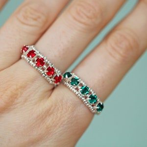 May include: Two silver rings with red and green gemstones. The rings are on a person's finger.