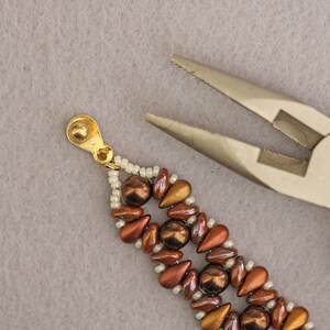 Peut inclure: Gros plan sur un bracelet en perles brunes et dor&eacute;es avec des perles de rocaille blanches. Le bracelet est tenu par une pince.
