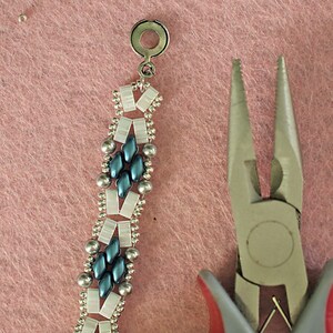 Op de afbeelding: Close-up van een zilveren en blauwe kralenarmband met een zilveren sluiting. Een tang bevindt zich op de voorgrond.