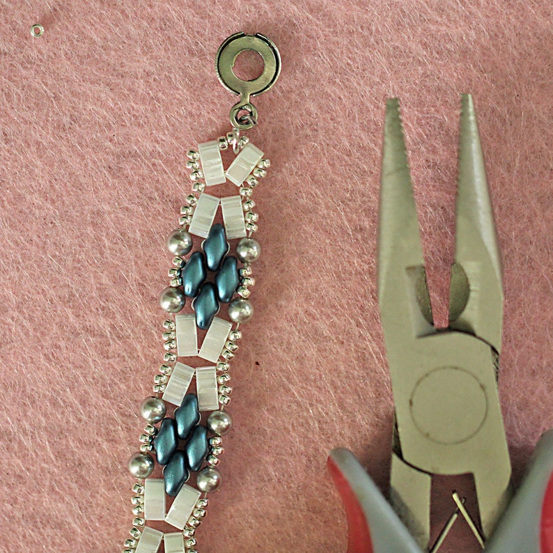 Op de afbeelding: Close-up van een zilveren en blauwe kralenarmband met een zilveren sluiting. Een tang bevindt zich op de voorgrond.