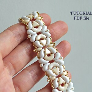 Op de afbeelding: Een witte en cr&egrave;mekleurige kralenarmband met een geometrisch patroon. De armband is gemaakt met kleine, ronde kralen en grotere, halvemaanvormige kralen. De kralen zijn geregen op een gouden draad. De armband wordt op een hand getoond, en de tekst "TUTORIAL PDF file" is zichtbaar in de rechterbovenhoek van de afbeelding.