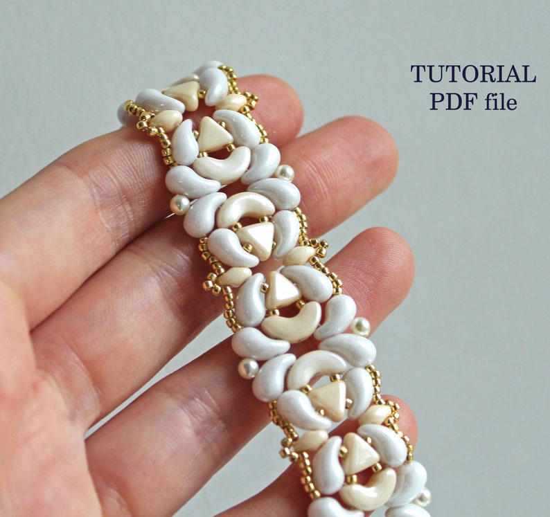 Op de afbeelding: Een witte en cr&egrave;mekleurige kralenarmband met een geometrisch patroon. De armband is gemaakt met kleine, ronde kralen en grotere, halvemaanvormige kralen. De kralen zijn geregen op een gouden draad. De armband wordt op een hand getoond, en de tekst "TUTORIAL PDF file" is zichtbaar in de rechterbovenhoek van de afbeelding.