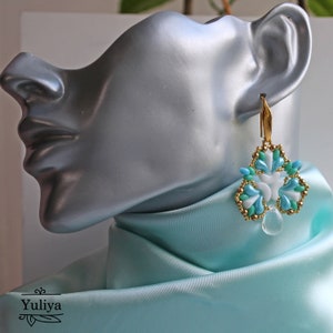 May include: A pair of dangle earrings with a floral design. The earrings are made with white and blue beads and gold accents. The earrings are hanging from a gold hook.