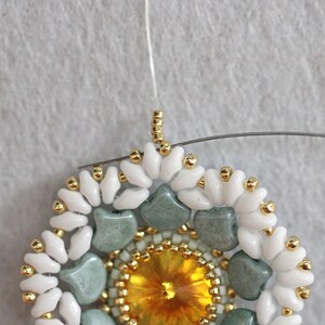 May include: Close-up of a beaded jewelry piece with a central yellow crystal surrounded by green and white beads. The piece is attached to a white thread.