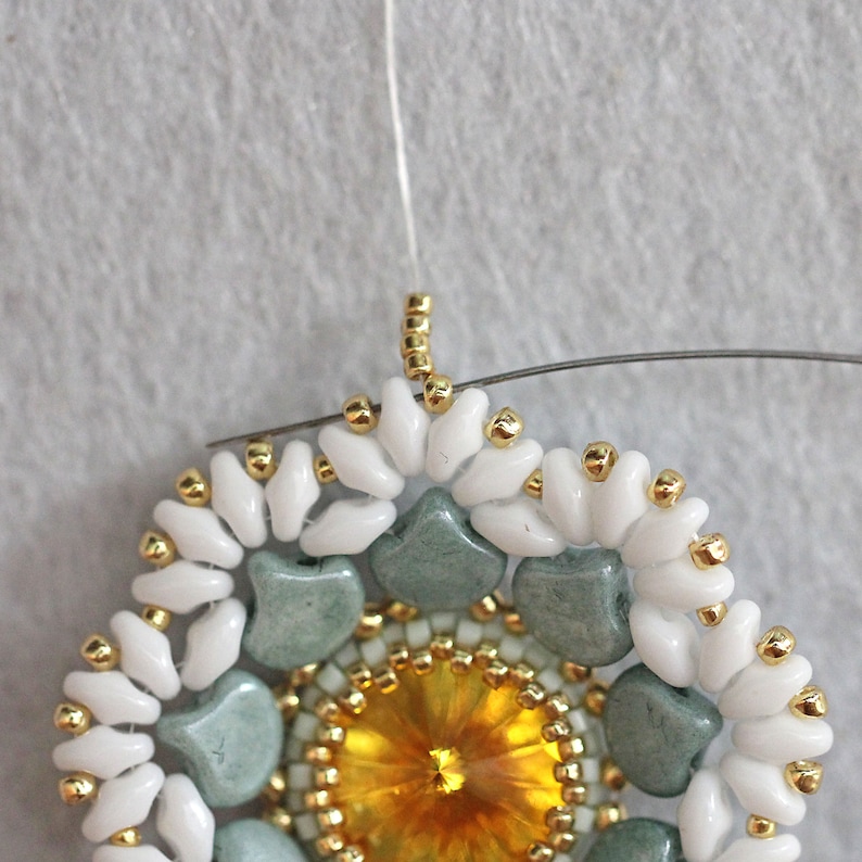 May include: Close-up of a beaded jewelry piece with a central yellow crystal surrounded by green and white beads. The piece is attached to a white thread.