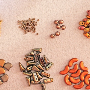 K&ouml;nnte beinhalten: Sortiment an bunten Glasperlen in verschiedenen Formen und Gr&ouml;&szlig;en, darunter runde, tropfenf&ouml;rmige und sichelf&ouml;rmige Formen. Die Perlen sind in Gruppen auf einer hellbeigen Oberfl&auml;che angeordnet.