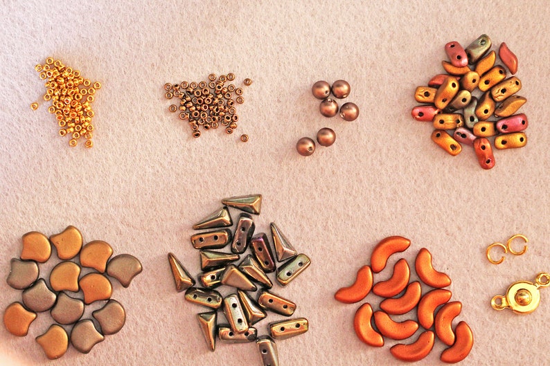 K&ouml;nnte beinhalten: Sortiment an bunten Glasperlen in verschiedenen Formen und Gr&ouml;&szlig;en, darunter runde, tropfenf&ouml;rmige und sichelf&ouml;rmige Formen. Die Perlen sind in Gruppen auf einer hellbeigen Oberfl&auml;che angeordnet.