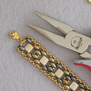 Puede incluir: Primer plano de un brazalete de cuentas doradas y plateadas con un cierre. El brazalete est&aacute; siendo sujetado por un par de alicates plateados con la palabra "Jewel" impresa en el lateral.