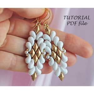 Può includere: Un paio di orecchini con perline bianche e dorate con un design geometrico. Gli orecchini sono appesi a ganci dorati. L'immagine include il testo "TUTORIAL PDF file".