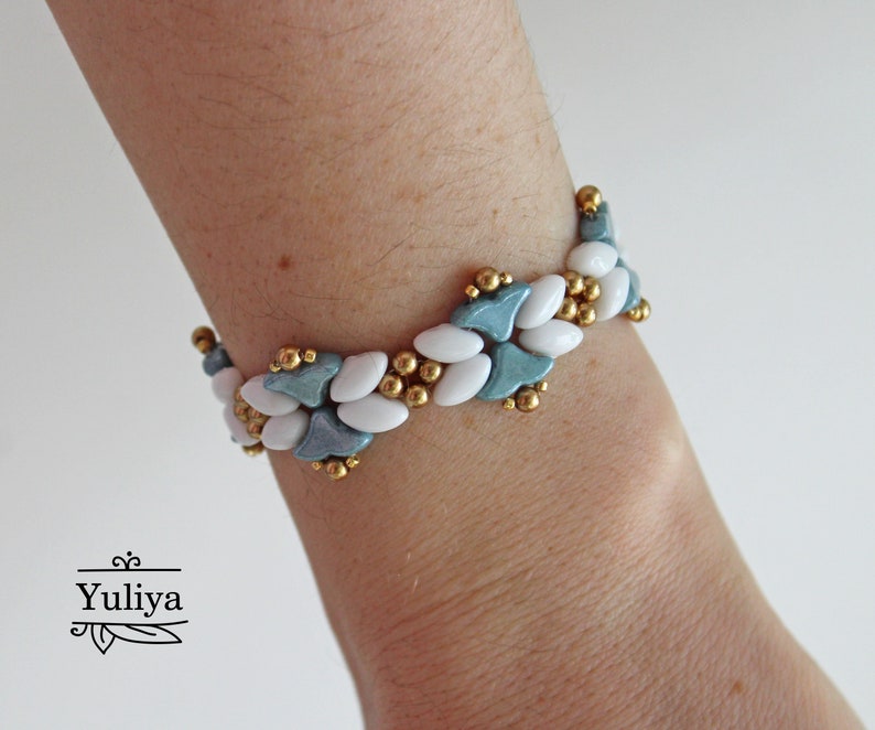 May include: A white and blue beaded bracelet with gold accents. The bracelet is made with small, oval-shaped beads and features a repeating pattern of blue and white.