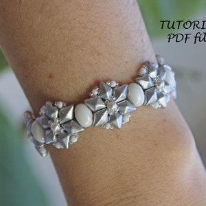 Puede incluir: Un brazalete de cuentas plateadas y blancas con un dise&ntilde;o de flores. El brazalete est&aacute; en la mu&ntilde;eca de una persona.  "TUTORIAL PDF file" est&aacute; escrito en la esquina superior derecha de la imagen.