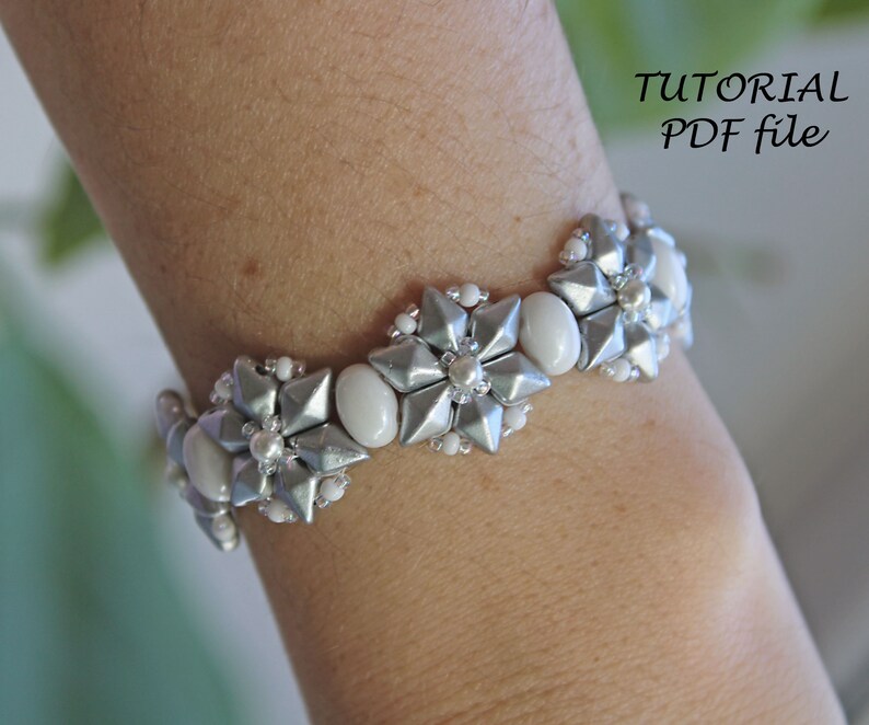 Puede incluir: Un brazalete de cuentas plateadas y blancas con un dise&ntilde;o de flores. El brazalete est&aacute; en la mu&ntilde;eca de una persona.  "TUTORIAL PDF file" est&aacute; escrito en la esquina superior derecha de la imagen.