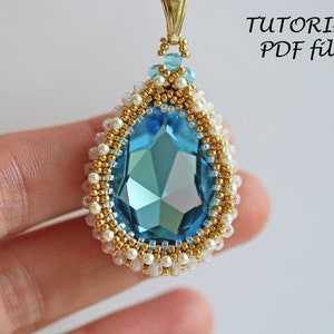 Peut inclure: Un pendentif en forme de larme avec une grande pierre pr&eacute;cieuse facet&eacute;e bleue entour&eacute;e de perles dor&eacute;es et blanches. Le pendentif est tenu par un doigt.