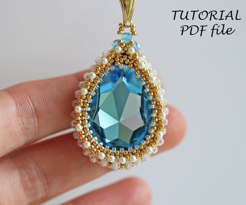 Peut inclure: Un pendentif en forme de larme avec une grande pierre pr&eacute;cieuse facet&eacute;e bleue entour&eacute;e de perles dor&eacute;es et blanches. Le pendentif est tenu par un doigt.