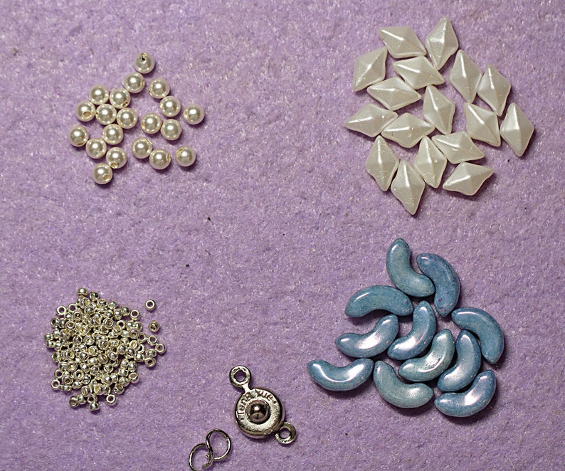 May include: Assortment of white, blue, and silver beads and findings. The white beads are in the shape of diamonds, the blue beads are crescent shaped, and the silver beads are round. There are also silver jump rings and a silver clasp.