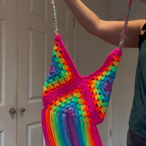 Puede incluir: Un bolso de crochet arcoíris con una larga correa trenzada y detalles de flecos. El bolso está hecho con colores brillantes, incluyendo rosa, naranja, amarillo, verde, azul y morado.