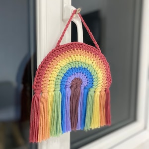 Könnte beinhalten: Ein regenbogenfarbener Wandbehang aus Häkelarbeit mit Quasten. Der Regenbogen besteht aus rotem, orangefarbenem, gelbem, grünem, blauem, indigoblauem und violettem Garn. Die Quasten haben die gleichen Farben wie der Regenbogen.