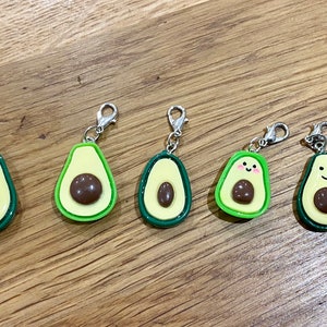 May include: Five avocado-shaped keyrings with silver clasps. The avocados are green and yellow with brown pits. Two of the avocados have a smiley face drawn on them.