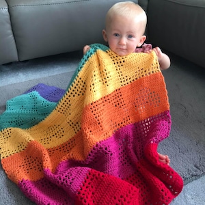 May include: A baby is sitting on a gray carpet holding a rainbow crocheted blanket. The blanket has stripes of teal, yellow, orange, pink, and red.