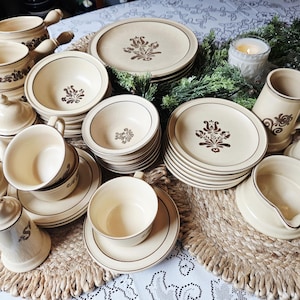 Puede incluir: Un juego de vajilla de cerámica beige con diseños florales marrones. El juego incluye platos, cuencos, tazas, platillos, tazas, una crema, un azucarero y saleros y pimenteros. Los platos están dispuestos sobre un mantel individual tejido.