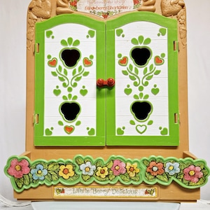 May include: A green and brown toy cabinet with a white door featuring a strawberry design. The cabinet has a shelf with a floral design and the text "Life is Berry Delicious".