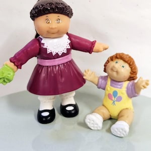 May include: Two Cabbage Patch Kids dolls. One doll is standing and wearing a burgundy dress, white collar, and black shoes. The other doll is sitting and wearing a yellow jumpsuit with a balloon graphic. Both dolls have curly hair.