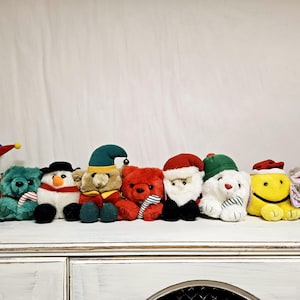 Peut inclure: Une collection de huit peluches habillées de costumes de fête. Les peluches comprennent un bonhomme de neige, un ours, un renne, un pingouin, un Père Noël, un chat et deux autres ours. Les peluches portent toutes des chapeaux et des écharpes.