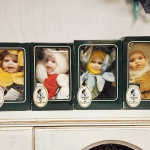 May include: Four vintage dolls in boxes, each with a unique outfit. The dolls have porcelain faces and are dressed in flower, fur, and other themed costumes. The boxes are dark green with a clear window and a logo.