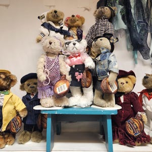 Könnte beinhalten: Eine Sammlung von Teddybären in verschiedenen Outfits, darunter eine gelbe Jacke, ein Paillettenkleid und Jeans. Die Bären stehen auf einem kleinen blauen Tisch und sind in verschiedenen Größen und Farben.