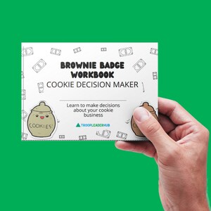 K&ouml;nnte beinhalten: Eine wei&szlig;e Karte mit dem Text "Brownie Badge Workbook Cookie Decision Maker" und einer Comic-Keksdose mit dem Text "Cookies!" darauf. Die Karte wird von einer Hand gehalten.