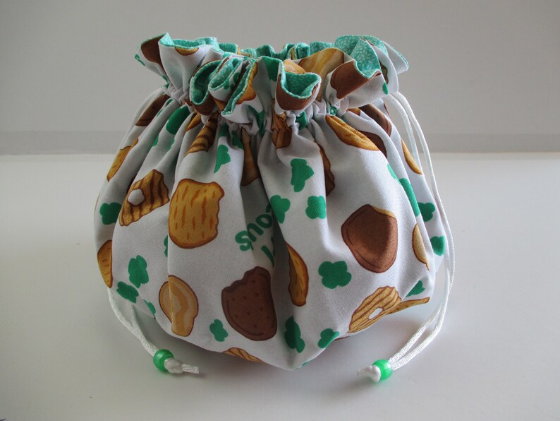 May include: White drawstring bag with a green and white print of Girl Scout cookies. The bag has a white drawstring with green beads.
