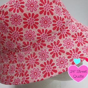 May include: Pink and red floral print bucket hat with a white brim. The hat has a label that says "24th Street Quilts".