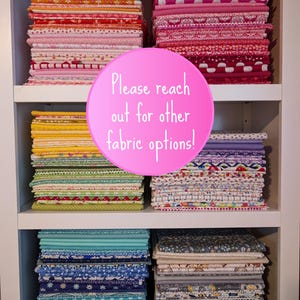 May include: A white shelf unit with multiple stacks of colorful fabric. The fabric is patterned with various designs, including polka dots, stripes, and floral prints. A pink circle with the text "Please reach out for other fabric options!" is placed in front of the shelf unit.