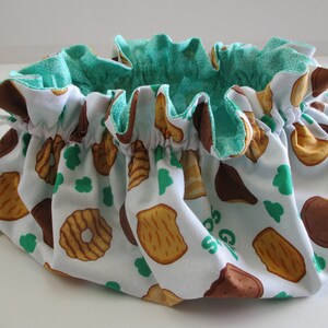 May include: A white drawstring bag with a green lining. The bag is decorated with a pattern of brown and tan cookies. The text "Girl Scouts" is visible on the bag.