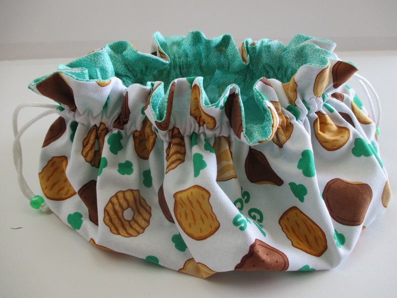 May include: A white drawstring bag with a green lining. The bag is decorated with a pattern of brown and tan cookies. The text "Girl Scouts" is visible on the bag.