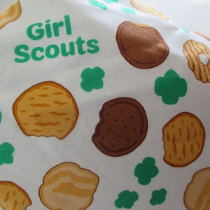 May include: White fabric with a green and brown pattern of Girl Scout cookies and four-leaf clovers. The text "Girl Scouts" is printed in green.