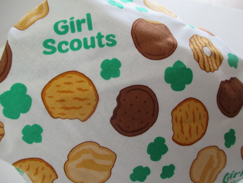May include: White fabric with a green and brown pattern of Girl Scout cookies and four-leaf clovers. The text "Girl Scouts" is printed in green.