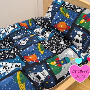 Può includere: Un set di trapunte e cuscini a tema spaziale. La biancheria da letto presenta un motivo ripetuto di razzi, pianeti e stelle su uno sfondo blu e nero. Il cuscino e la trapunta sono esposti su una cornice di legno. Il testo "24th Street Quilts" è visibile.