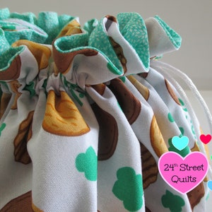 May include: A white drawstring bag with a green and brown print of cookies and shamrocks. The bag has a white drawstring and is tied at the top. The bag has a pink heart-shaped tag with the text "24th Street Quilts".
