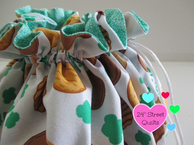 May include: A white drawstring bag with a green and brown print of cookies and shamrocks. The bag has a white drawstring and is tied at the top. The bag has a pink heart-shaped tag with the text "24th Street Quilts".